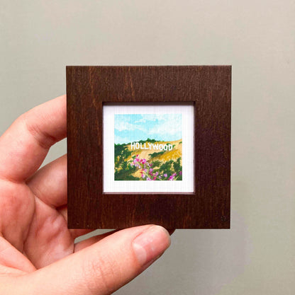 A hand is holding a small, square-shaped picture frame containing a painting of Hollywood.