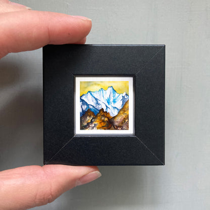 A person's hand holding a small, square-shaped black frame containing a watercolor painting of a mountain landscape.