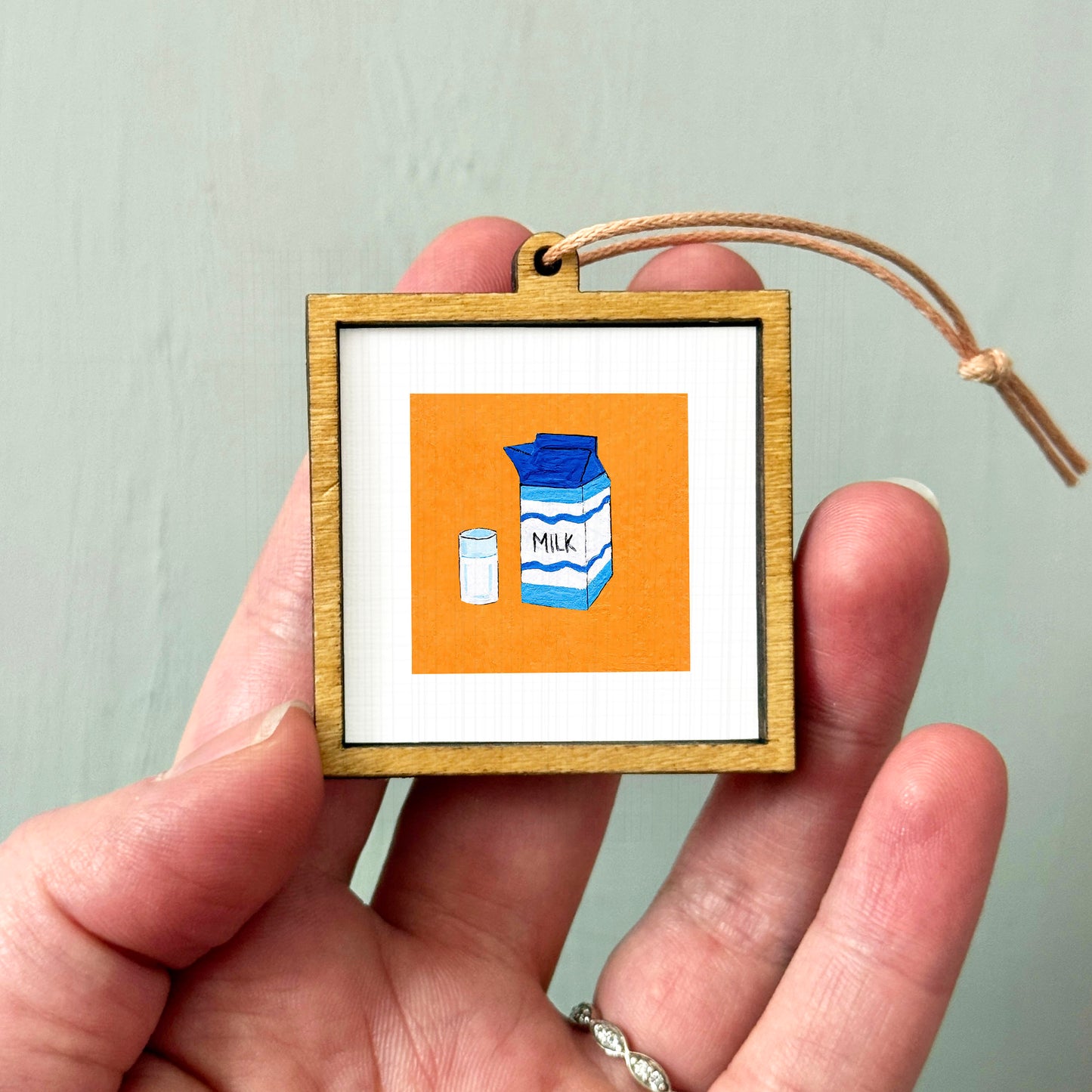 A person's hand holding a small wooden frame containing an illustration of a carton of milk and a glass of milk.