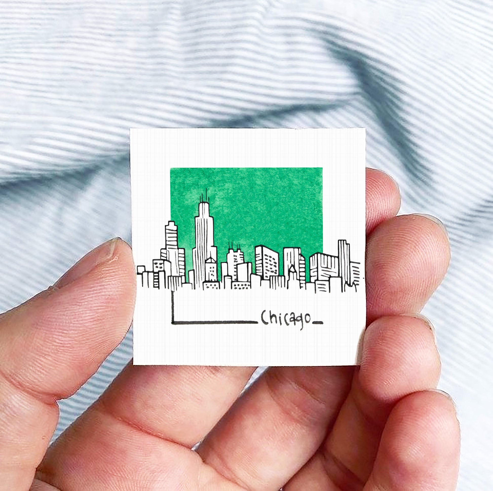 A hand is holding a small square-shaped object with a green background depicting a city skyline and the word "CHICAGO" written below it.
