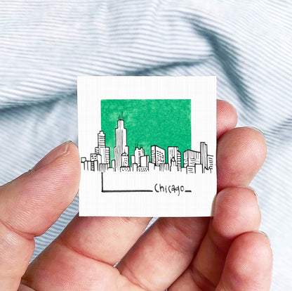A hand is holding a small square-shaped object with a green background depicting a city skyline and the word "CHICAGO" written below it.