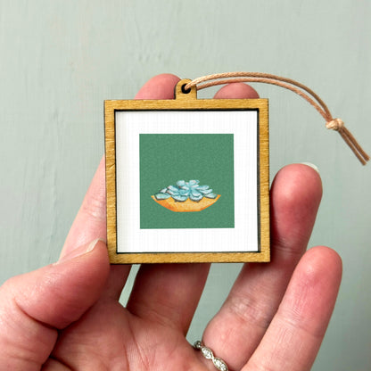 A hand holding a small wooden frame with a picture of a green plant inside it.