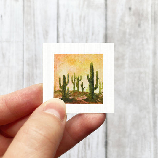 A hand is holding a small square-shaped picture of a desert scene with cacti.
