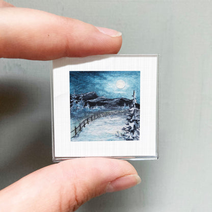 A person's hand holding a small square picture depicting a snowy landscape with a full moon in the night sky.