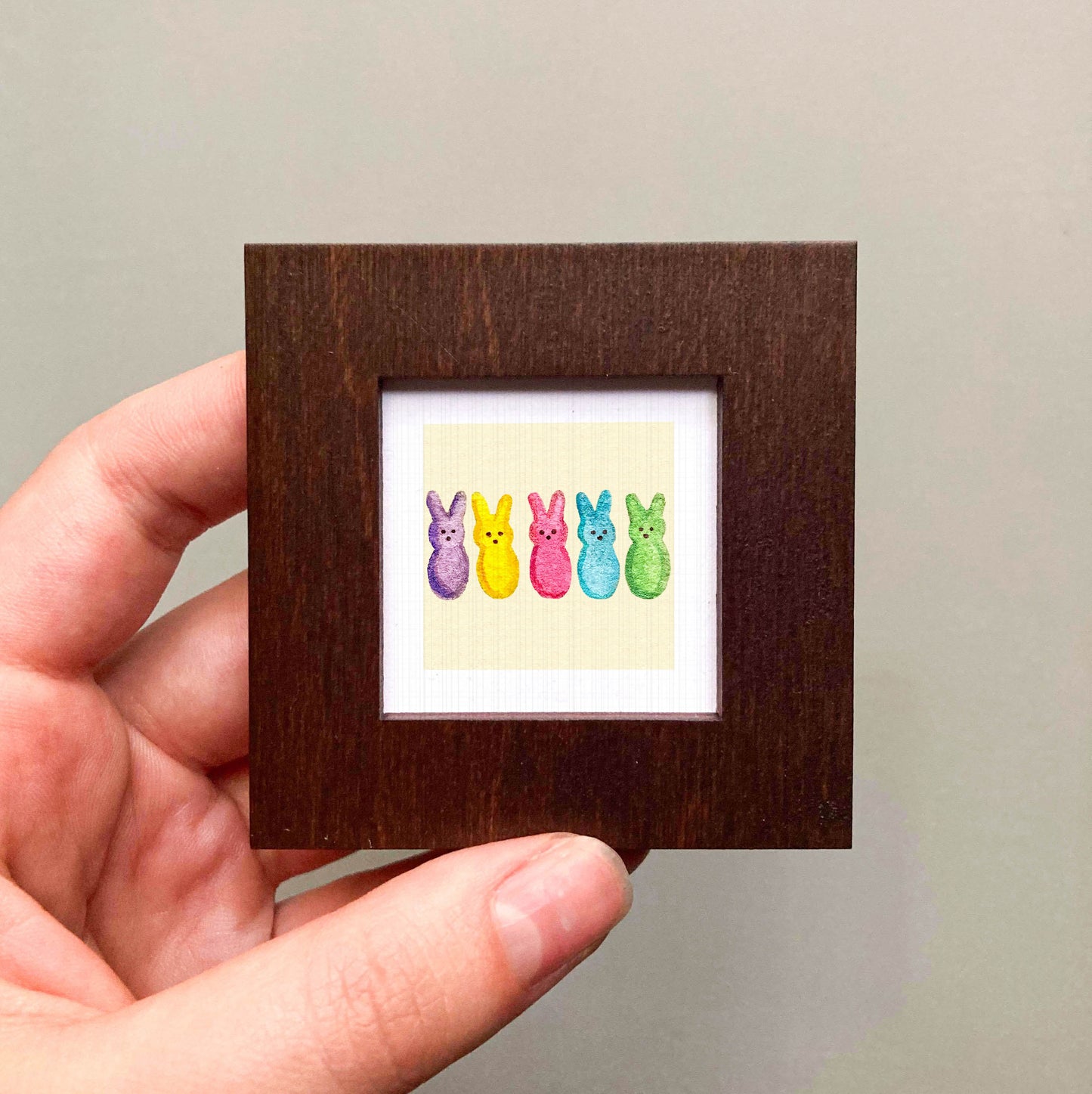 A hand is holding a small wooden frame containing a colorful illustration of five Easter bunny characters in various pastel shades.