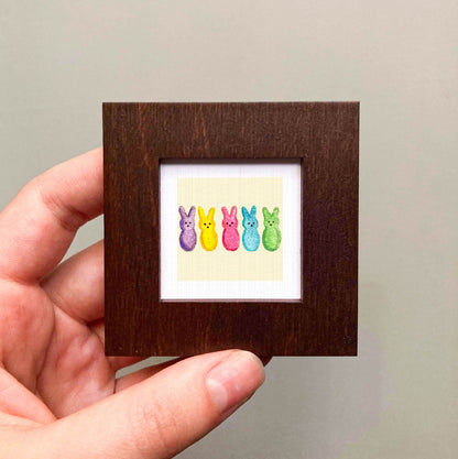 A hand is holding a small wooden frame containing a colorful illustration of five Easter bunny characters in various pastel shades.