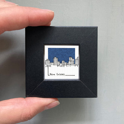 A hand is holding a small black frame containing a framed illustration of a city skyline.