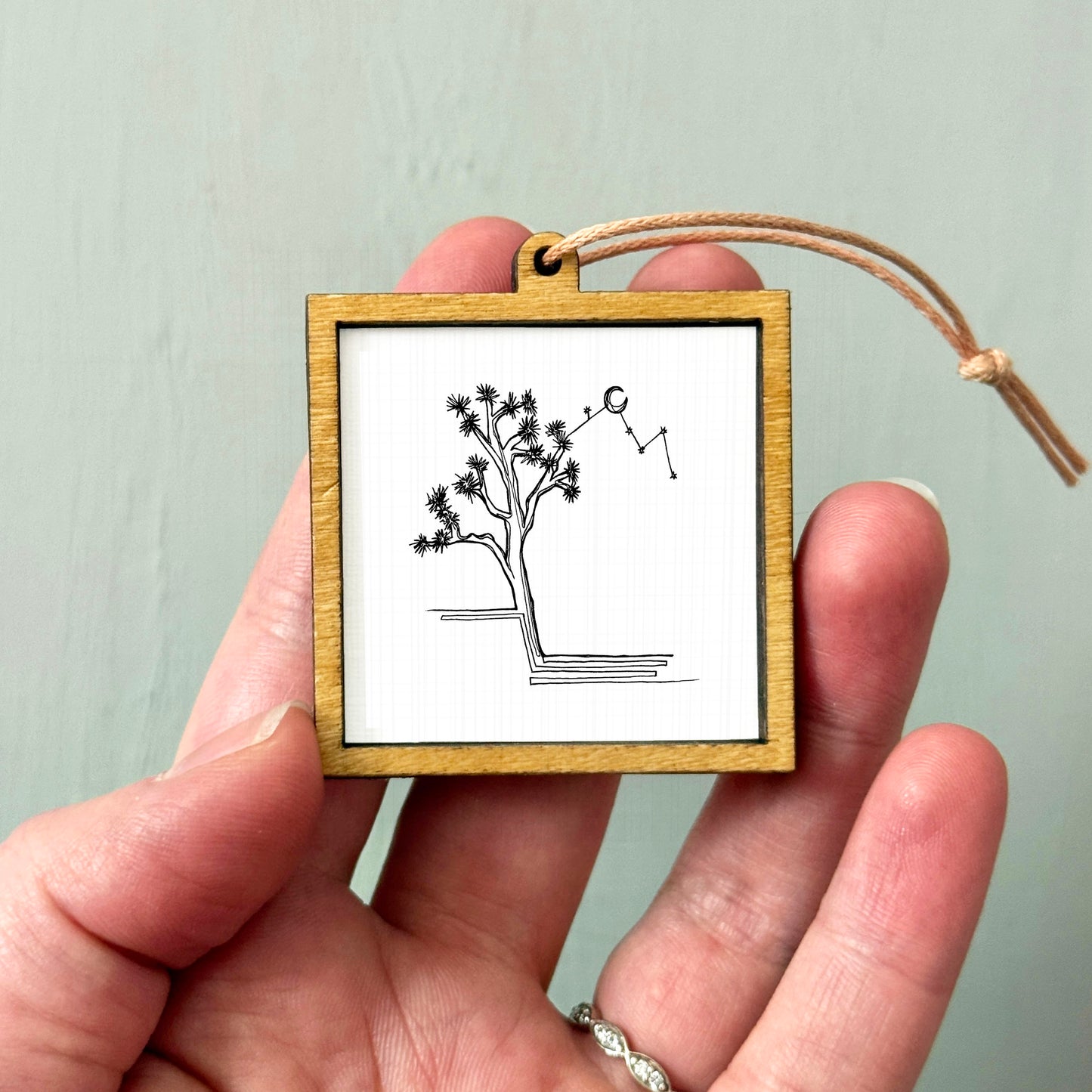 A hand holding a small wooden frame containing a simple black and white line drawing of a tree.