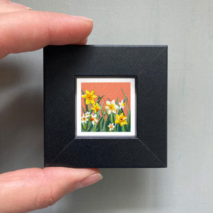 A person's hand holding a small black frame containing a framed painting of yellow and white flowers.