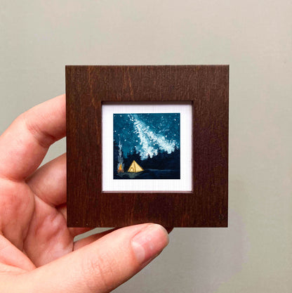 A hand is holding a small wooden frame containing a framed painting of a starry night sky with a tent in the foreground.