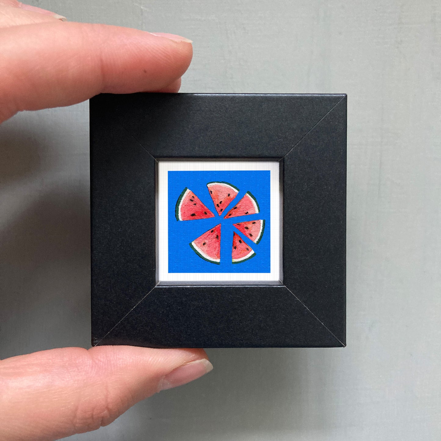 A hand is holding a small black square frame containing a vibrant illustration of watermelon slices against a blue background.