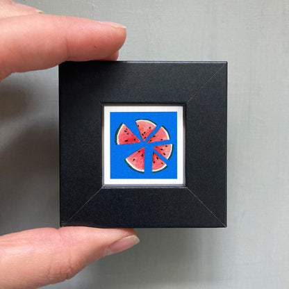 A hand is holding a small black square frame containing a vibrant illustration of watermelon slices against a blue background.