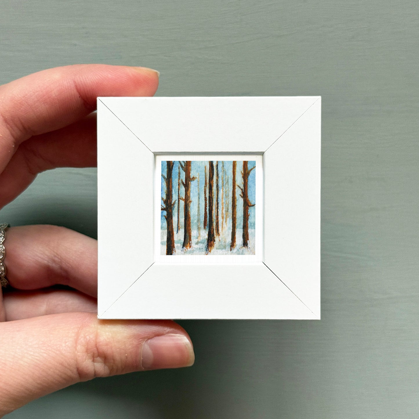 A hand is holding a small white square frame containing a painting of trees in a snowy landscape.