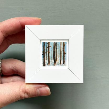 A hand is holding a small white square frame containing a painting of trees in a snowy landscape.