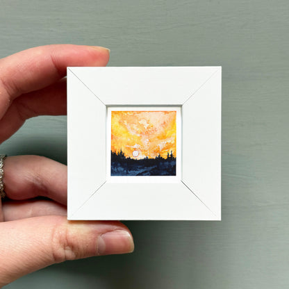 A hand is holding a small white square frame containing a painting of a sunset over a forest.