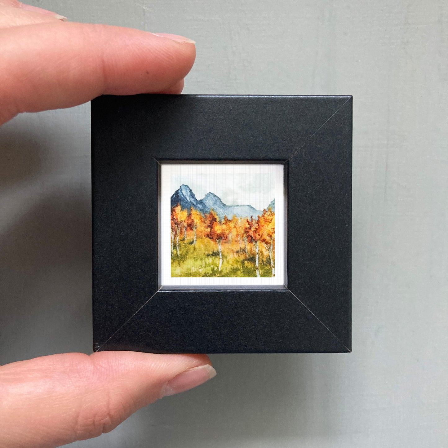 A person's hand holding a small, square-shaped black frame containing a watercolor painting of a mountain landscape with autumn trees.