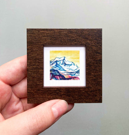 A hand is holding a small, square-shaped wooden frame containing a colorful painting of a mountain landscape.