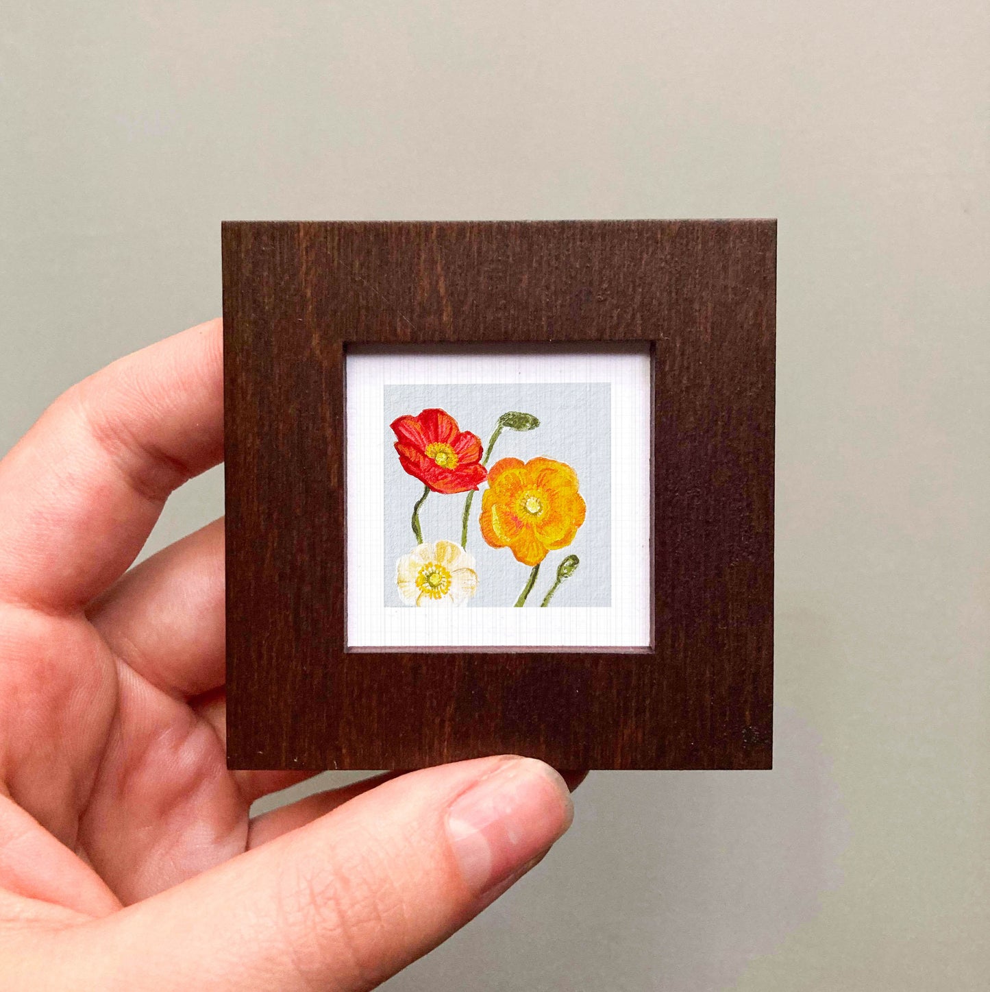 A hand is holding a small wooden frame containing a framed painting of two flowers, one red and one yellow.