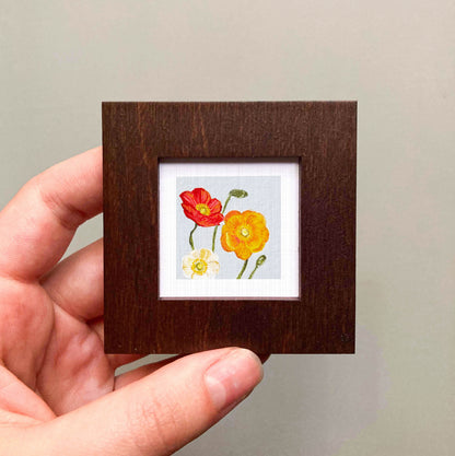 A hand is holding a small wooden frame containing a framed painting of two flowers, one red and one yellow.