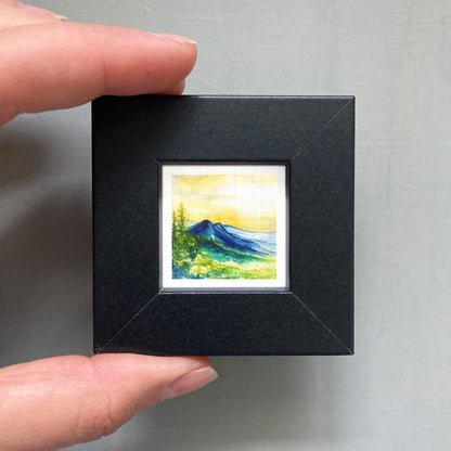 A person's hand holding a small, square-shaped black frame containing a watercolor painting of a mountain landscape.