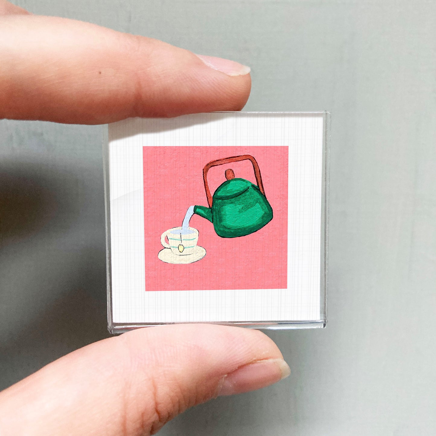 A person's hand holding a small square with a green teapot and a cup on a pink background.