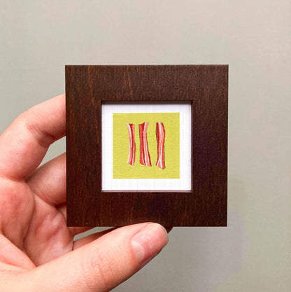 A hand is holding a small square frame containing a framed artwork of three red and white striped objects.