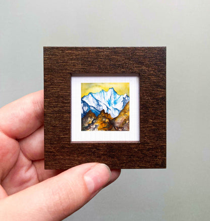 A hand is holding a small, square-shaped wooden frame containing a watercolor painting of a mountain landscape.