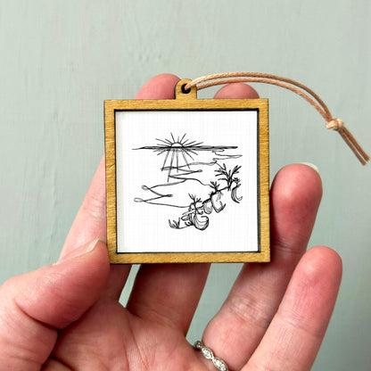 A hand holding a small wooden frame with a black and white illustration of a landscape on it.