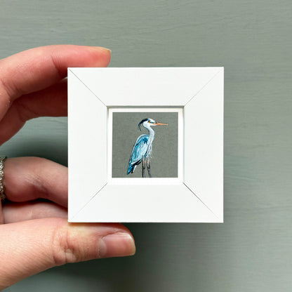 A hand is holding a small white square frame containing a painting of a blue heron.