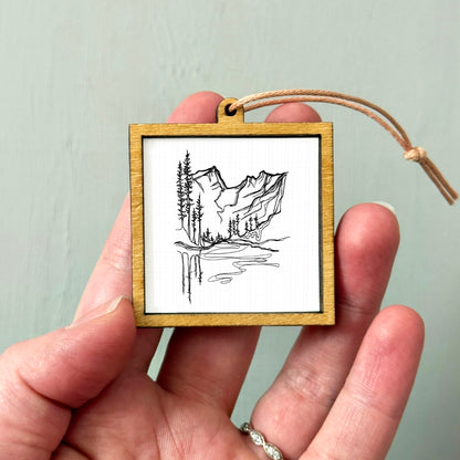 A hand holding a small wooden frame containing a black and white illustration of a mountain landscape.