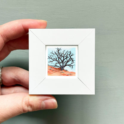 A hand is holding a small white square frame containing a painting of a bare tree with a red and orange background.