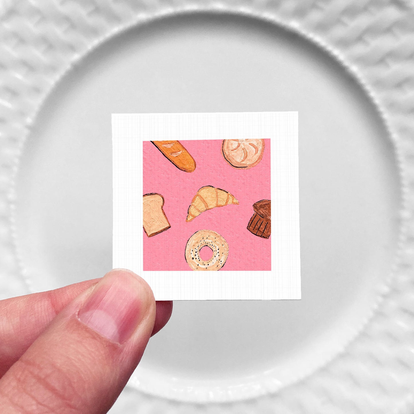 A hand is holding a small square with an illustration of various pastries, including a croissant, a bagel, and a doughnut, against a pink background.