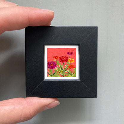 A hand is holding a small black frame containing a vibrant painting of colorful flowers against a red background.