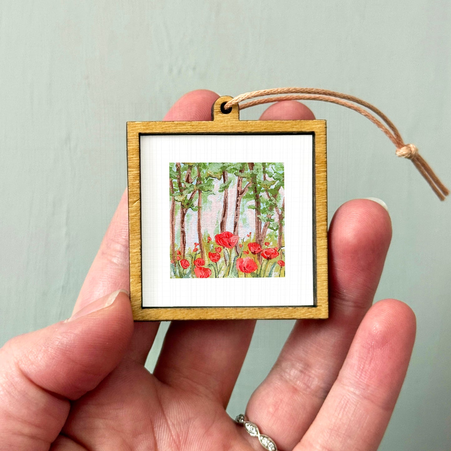 A hand holding a small wooden frame containing a painting of red flowers in a forest.