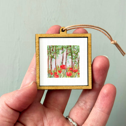 A hand holding a small wooden frame containing a painting of red flowers in a forest.