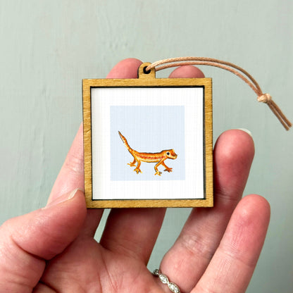 A person's hand holding a small wooden frame containing a picture of a gecko.