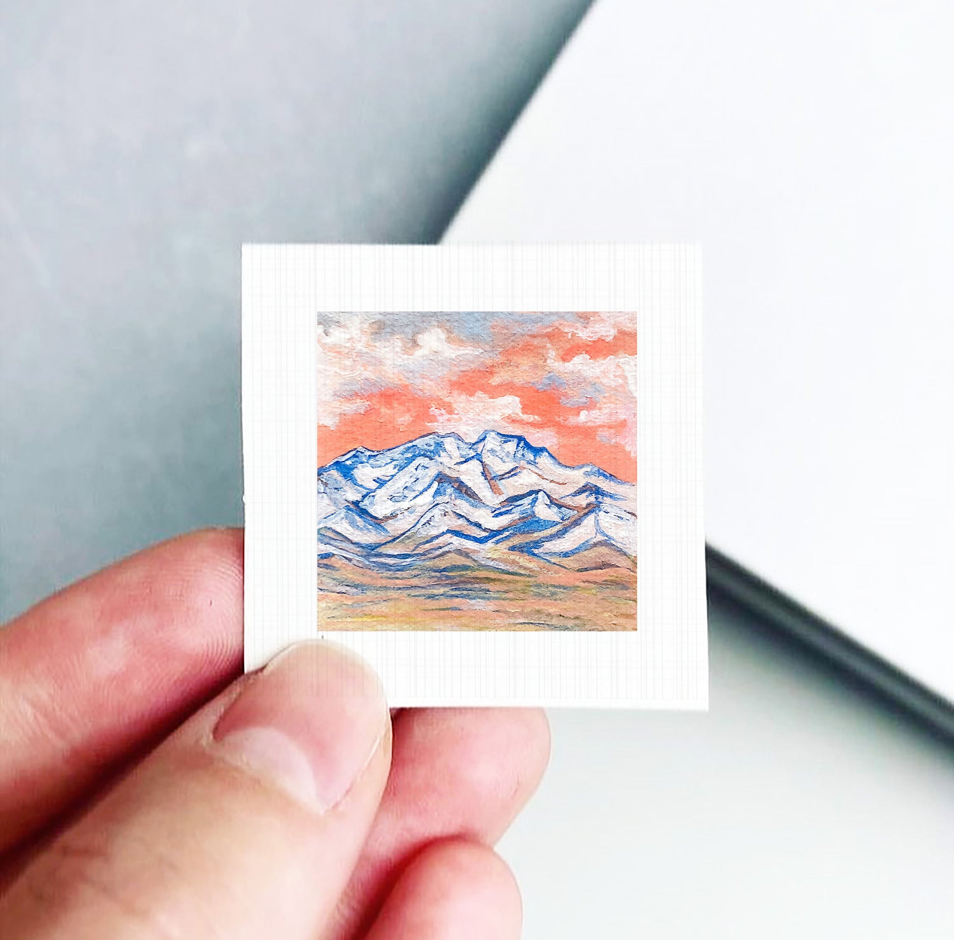 A hand is holding a small square picture of a mountain landscape with a colorful sky in the background.