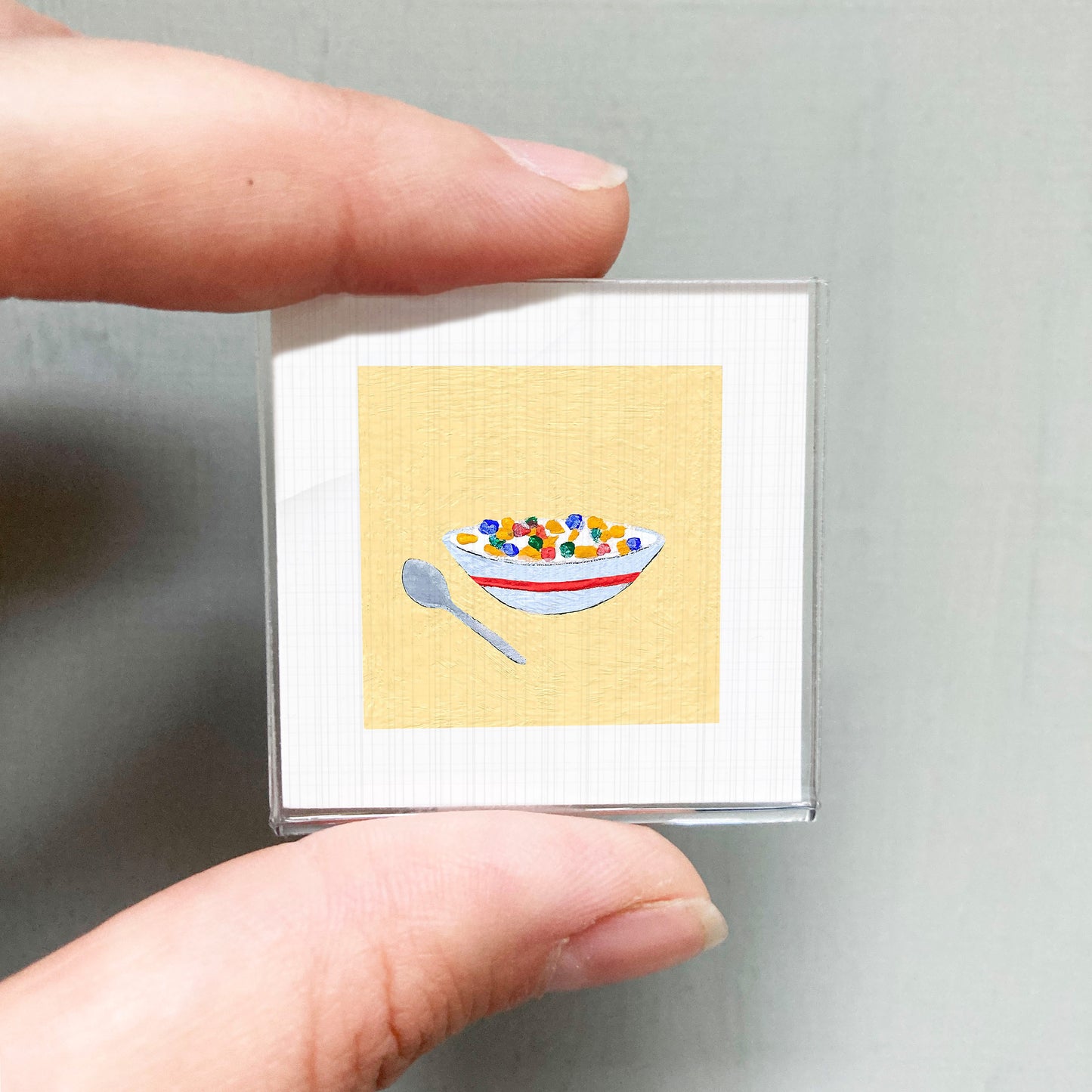 A person's hand holding a small square with a colorful bowl of cereal and a spoon on it.