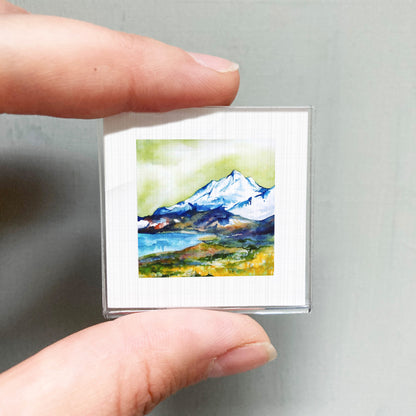 A person's hand is holding a small, square-shaped picture of a mountain landscape with a lake and green grass.