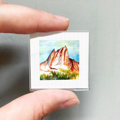 A person's hand holding a small square picture of a mountain landscape with a watercolor painting of the mountain.