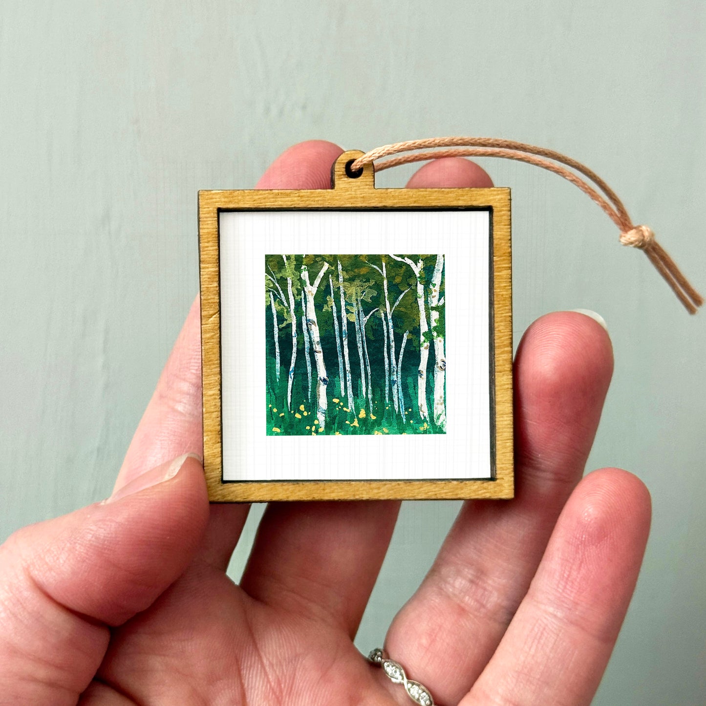 A hand holding a small wooden frame containing a painting of a forest scene with birch trees.