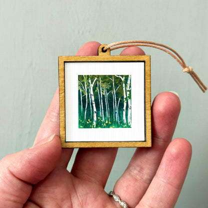 A hand holding a small wooden frame containing a painting of a forest scene with birch trees.