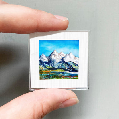 A person's hand holding a small framed painting of a mountain landscape.