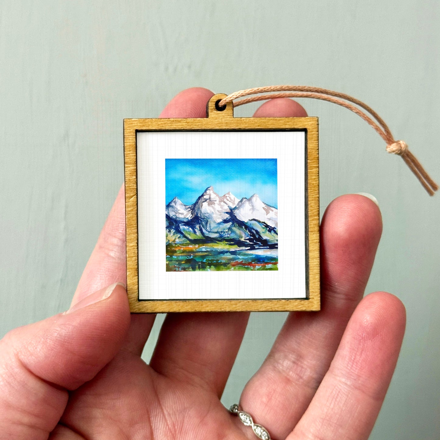 A person's hand holding a small wooden frame containing a painting of a mountain landscape.