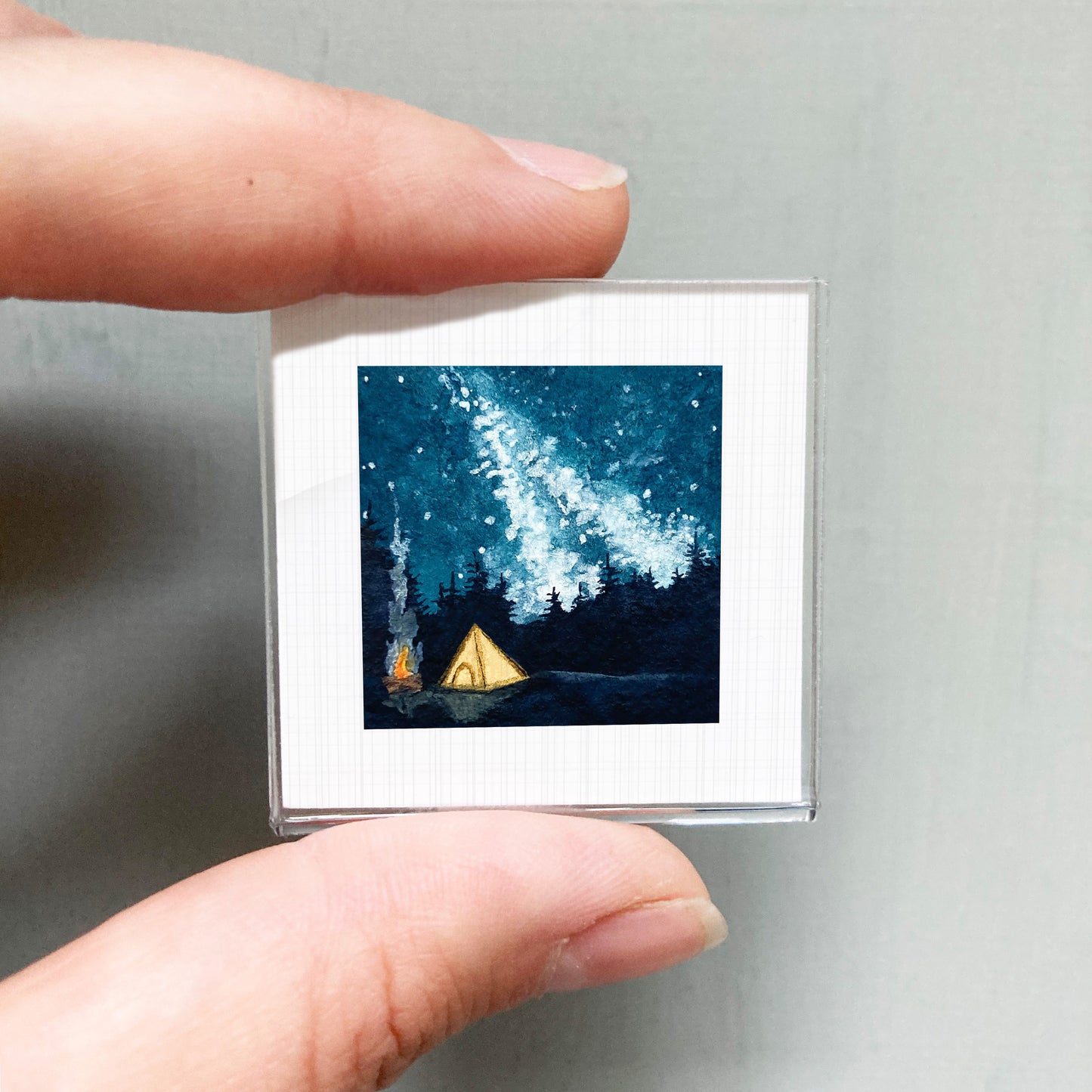 A person's hand is holding a small square picture depicting a nighttime scene with a tent, campfire, and a starry sky.