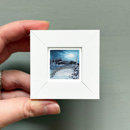 A hand is holding a small white square frame containing a painting of a snowy landscape with a full moon in the sky.