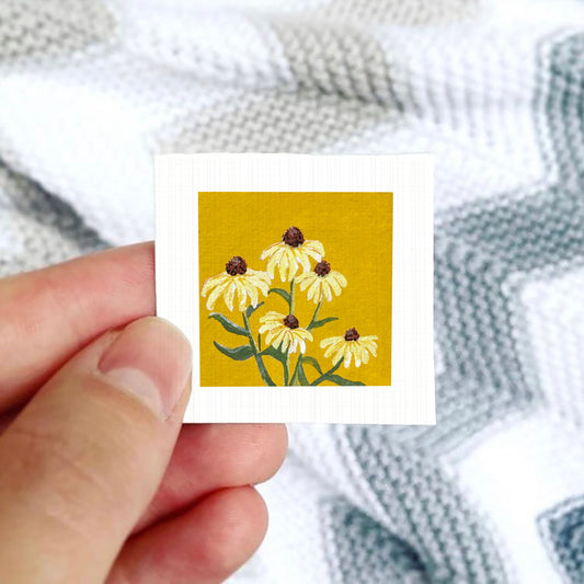 A hand is holding a small square card with a vibrant yellow flower design on it, set against a blurred background of a white and gray patterned fabric.