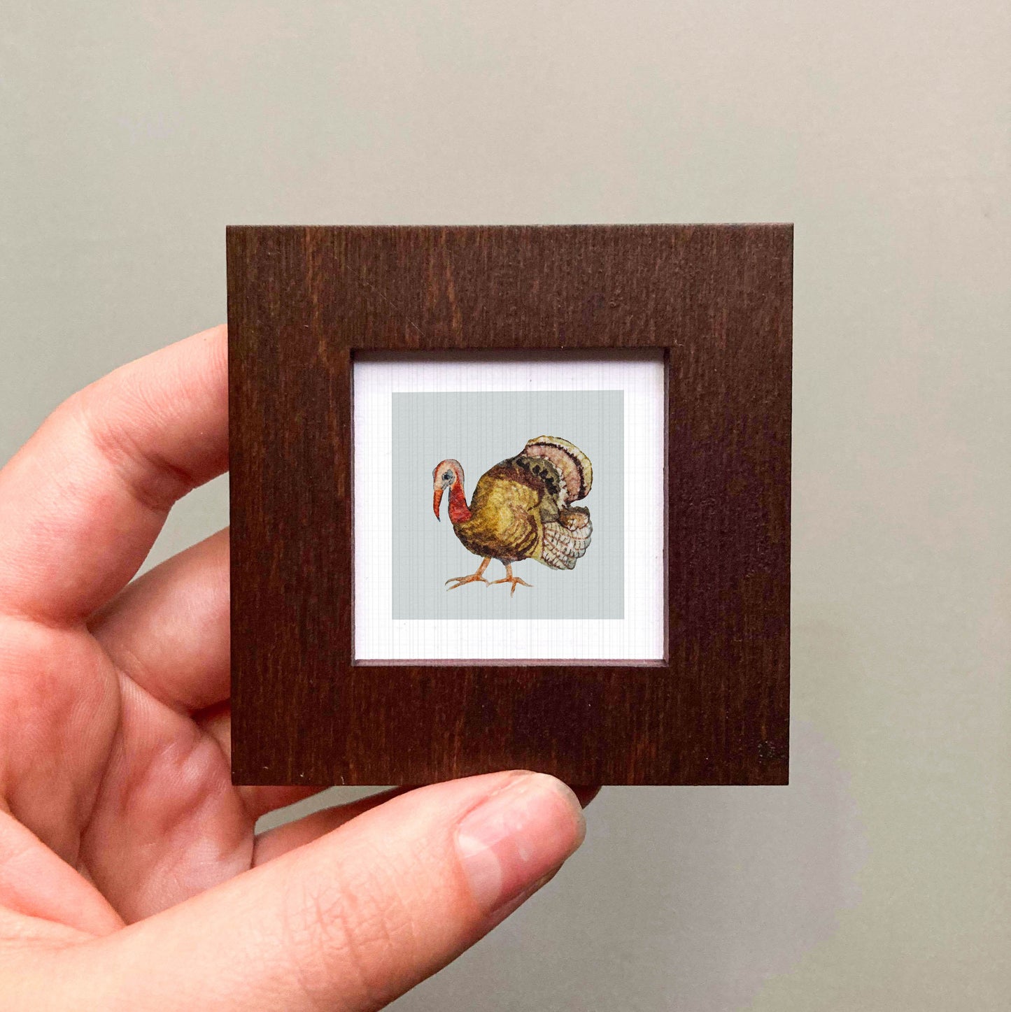 A hand is holding a small wooden frame containing a framed illustration of a turkey.