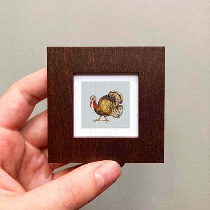 A hand is holding a small wooden frame containing a framed illustration of a turkey.