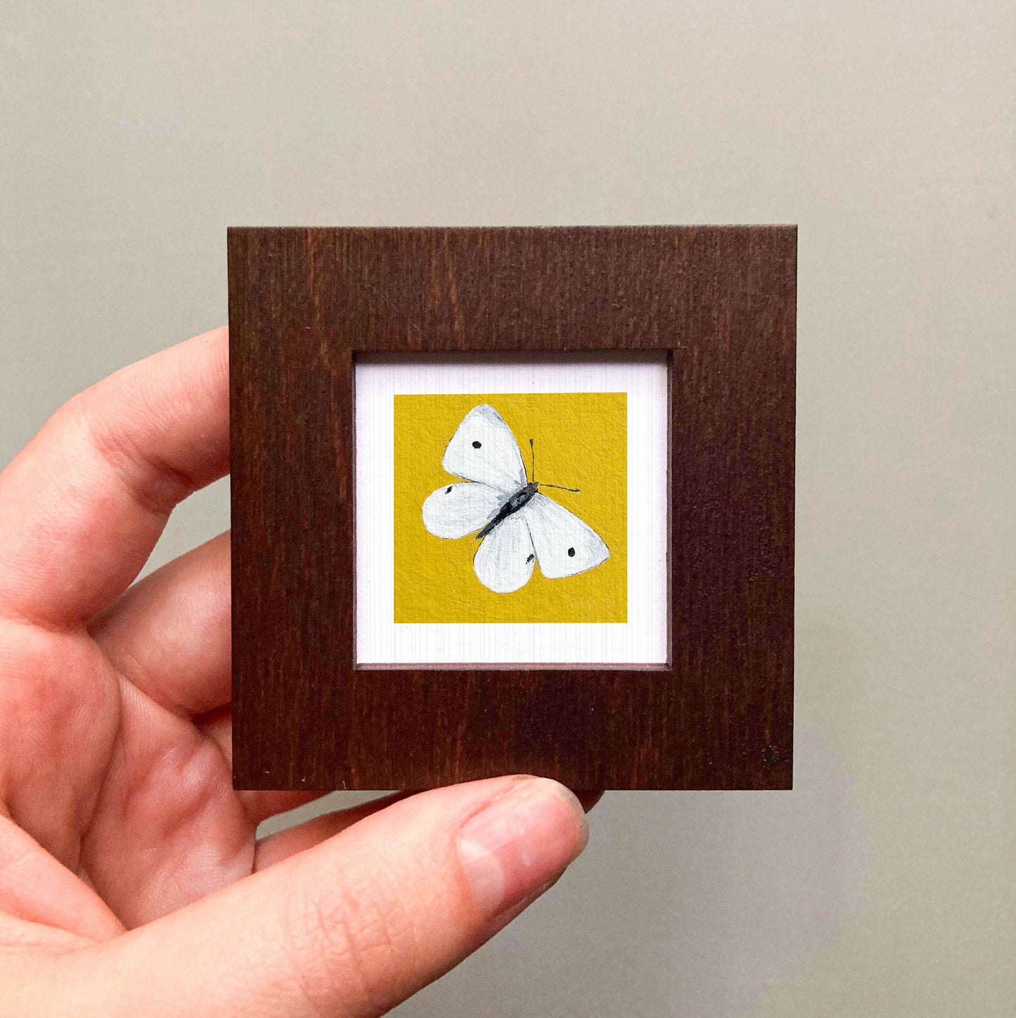 A hand is holding a small wooden frame containing a framed image of a white butterfly against a yellow background.